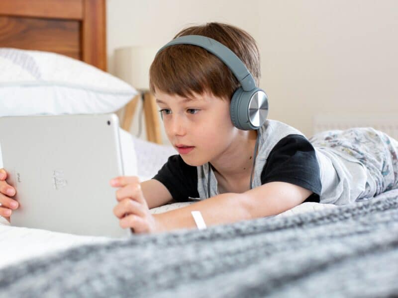 A young boy wearing headphones lying on a bed holding his tablet device.
