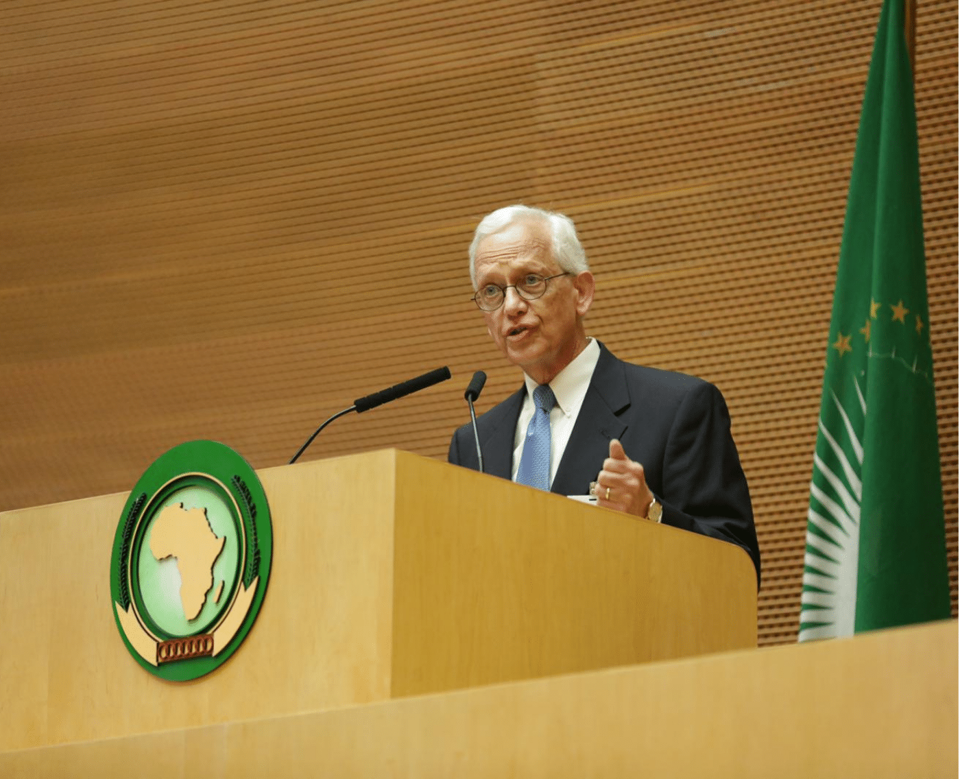 Ernie Allen, Chair of WePROTECT, is seen standing at a podium, delivering the opening remarks at the Global Summit to Tackle Online Sexual Exploitation. He is wearing a dark suit with a light blue tie and glasses. Behind him is a large wooden wall with a green African Union flag and a seal displaying the African continent. He is speaking into a microphone, with a serious and focused expression, highlighting the importance of the summit’s mission. 