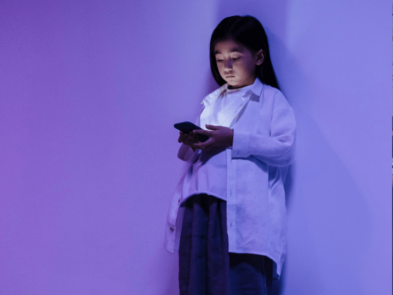 A young girl standing against a plain wall, bathed in soft purple and blue lighting. She is wearing a loose white shirt over a white top, paired with dark-coloured pants or a skirt. The girl is looking down intently at a smartphone that she is holding with both hands. The light from the phone softly illuminates her face.
