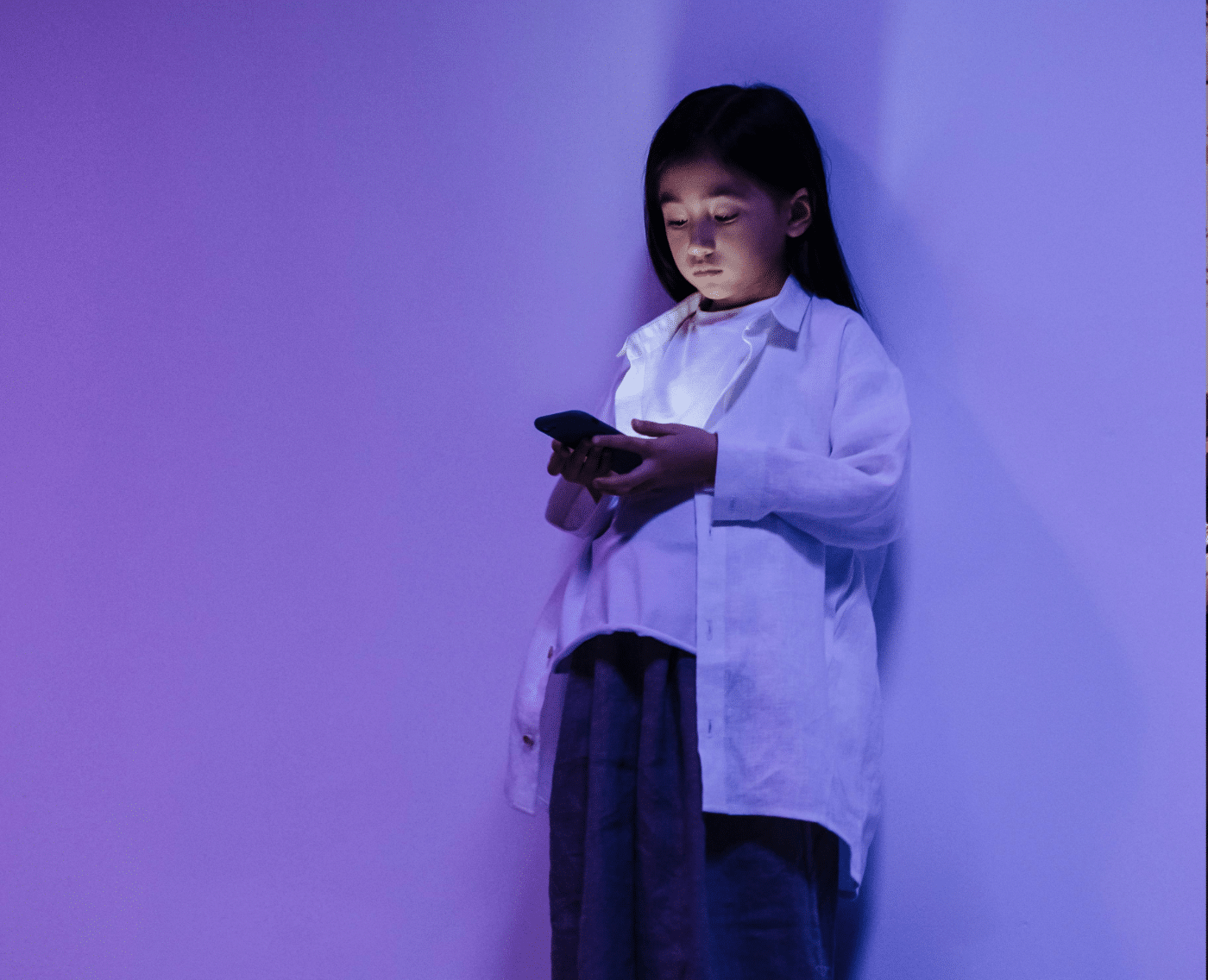 A young girl standing against a plain wall, bathed in soft purple and blue lighting. She is wearing a loose white shirt over a white top, paired with dark-coloured pants or a skirt. The girl is looking down intently at a smartphone that she is holding with both hands. The light from the phone softly illuminates her face.