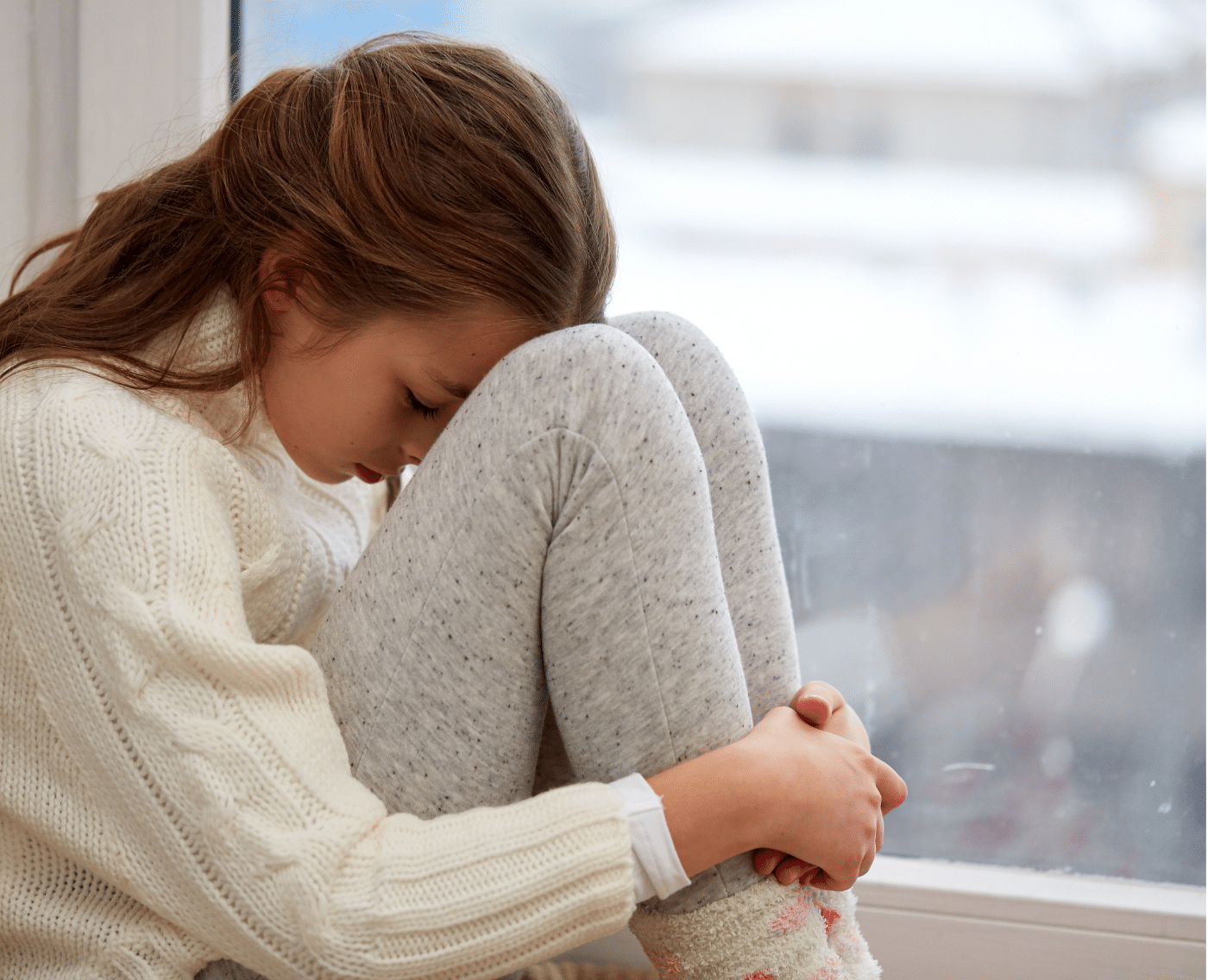 A full view of the young girl sitting in the same position by the window. The girl is hugging her knees, with her head resting on them.