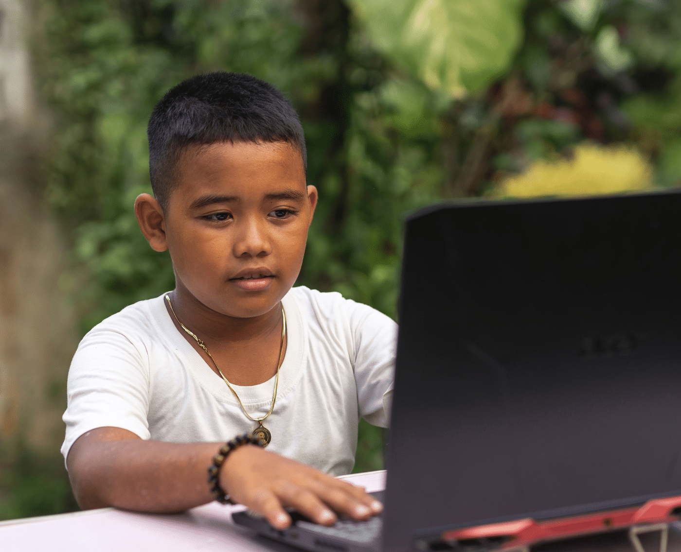 A young Asian boy on a laptop sat on a desk outside (you can see large foliage in the background that suggests this is in Southeast Asia). The camera is positioned so you can see the face of the boy looking at the laptop as he types. He looks inquisitive. 
