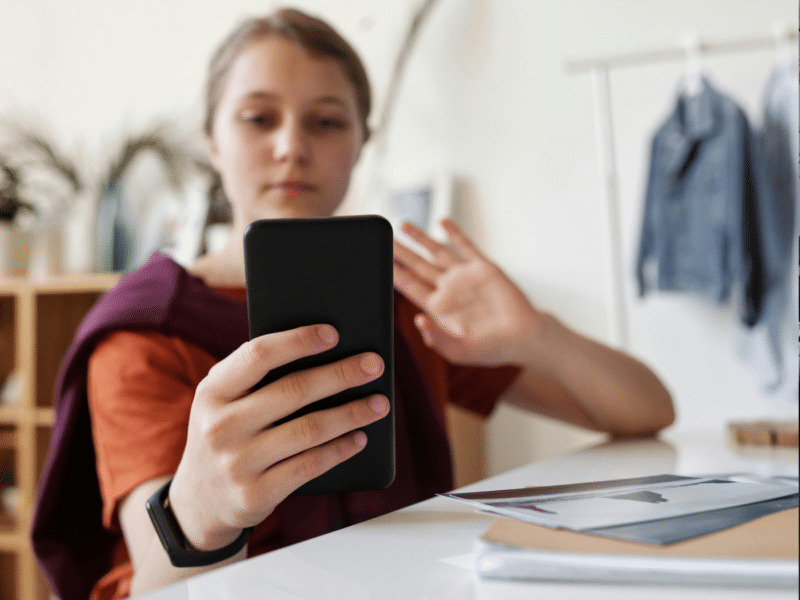 A young person is sitting indoors at a desk and holding a smartphone in their right hand. They appear to be engaged in a video call, as they are looking at the phone's screen and raising their left hand, possibly waving. The person is wearing an orange shirt and has a dark smartwatch on their left wrist. In the background, there is a shelf with potted plants and some hanging clothing, which is slightly out of focus, suggesting the photo was taken in a home or casual setting.
