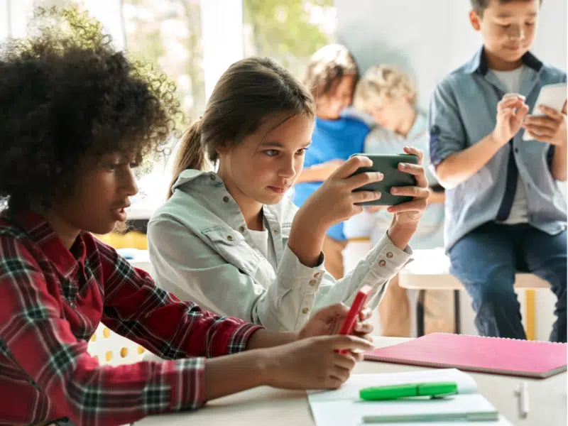 A classroom setting with a group of children engaging with digital technology. Two girls sit at a table in the foreground; one in a plaid shirt is holding a smartphone with a red cover, while the other in a white jacket is focused on her own smartphone. The table has school supplies, including a notebook and markers. In the background, three boys are sitting on a desk, interacting with tablets and smartphones. The scene is well-lit, with natural light streaming through large windows.