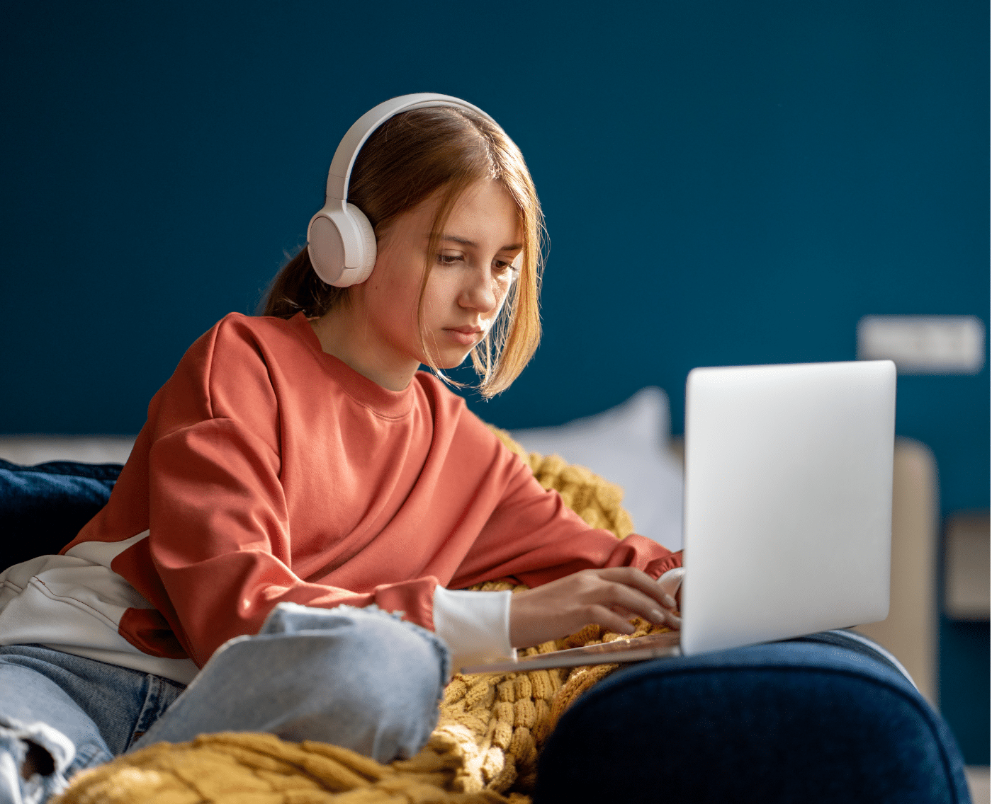 A wider shot of the same teenage girl seated comfortably on a couch at home. She is wearing a rust-orange sweatshirt, light blue ripped jeans, and white over-ear headphones, focused on her laptop. A yellow knitted blanket and blue wall add to the cozy setting. This image is a fuller version of the close-up shown above on the same page.