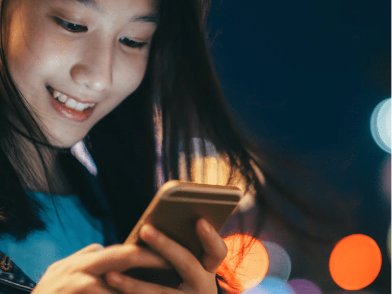 A wider shot of the same young person engrossed in their smartphone. The scene captures the person holding the phone with both hands, with the colorful city lights in the background. The individual is wearing a denim jacket, and their expression is one of joy. This is the full version of the close-up shown above.