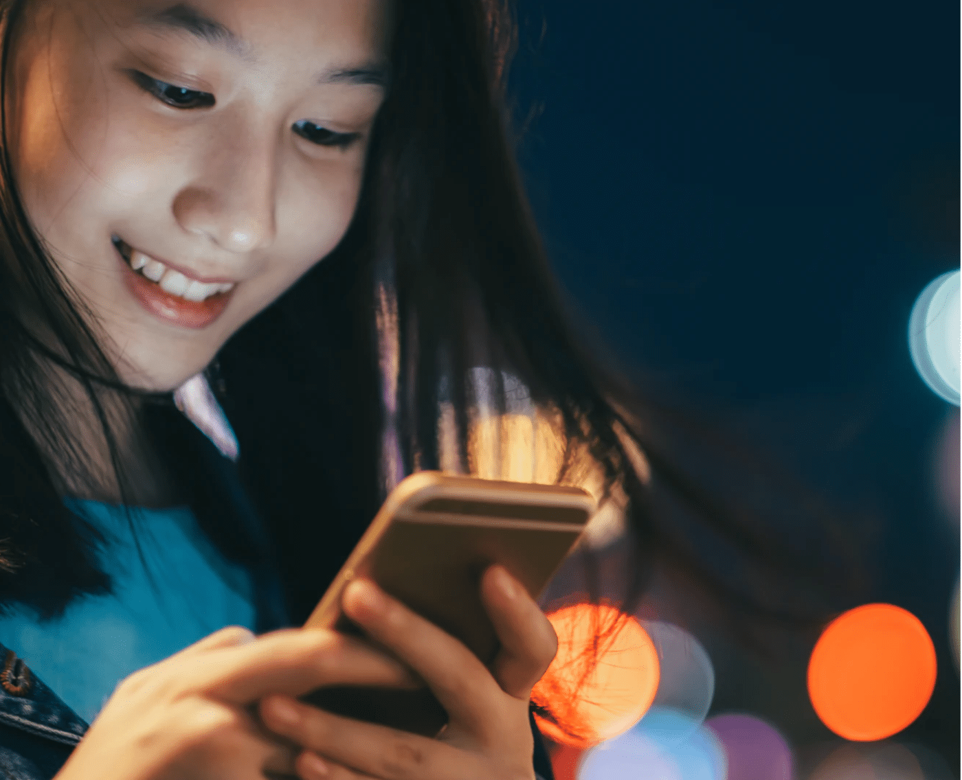 A wider shot of the same young person engrossed in their smartphone. The scene captures the person holding the phone with both hands, with the colorful city lights in the background. The individual is wearing a denim jacket, and their expression is one of joy. This is the full version of the close-up shown above.